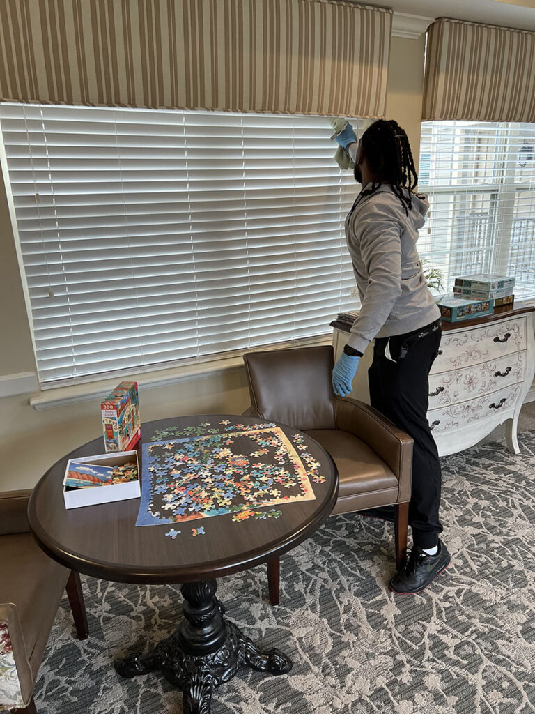 Staff members clean window blinds in a room with a puzzle on the table in the senior living community.