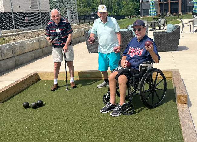 Three elderly men, one in a wheelchair, happily play bocce ball outside at Vitalia Mentor Senior Living Community.