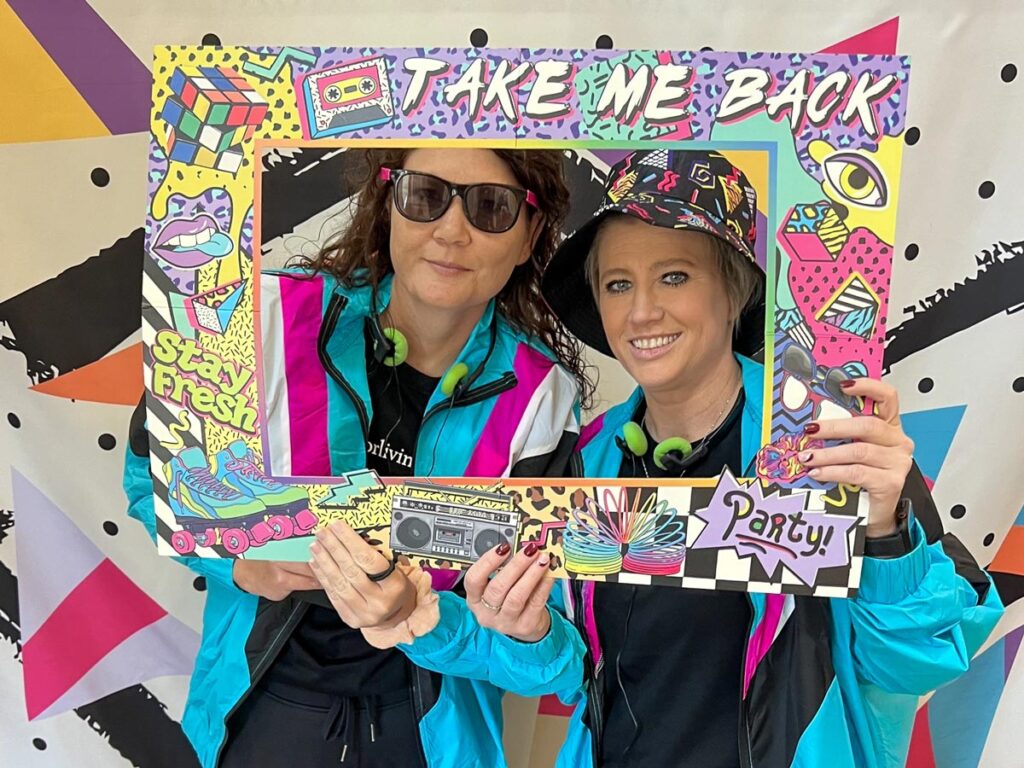 Two women at a job fair pose with a retro-themed 80s photo frame that says "Take Me Back," dressed in vibrant 80s outfits with neon colors, boombox, and playful designs.