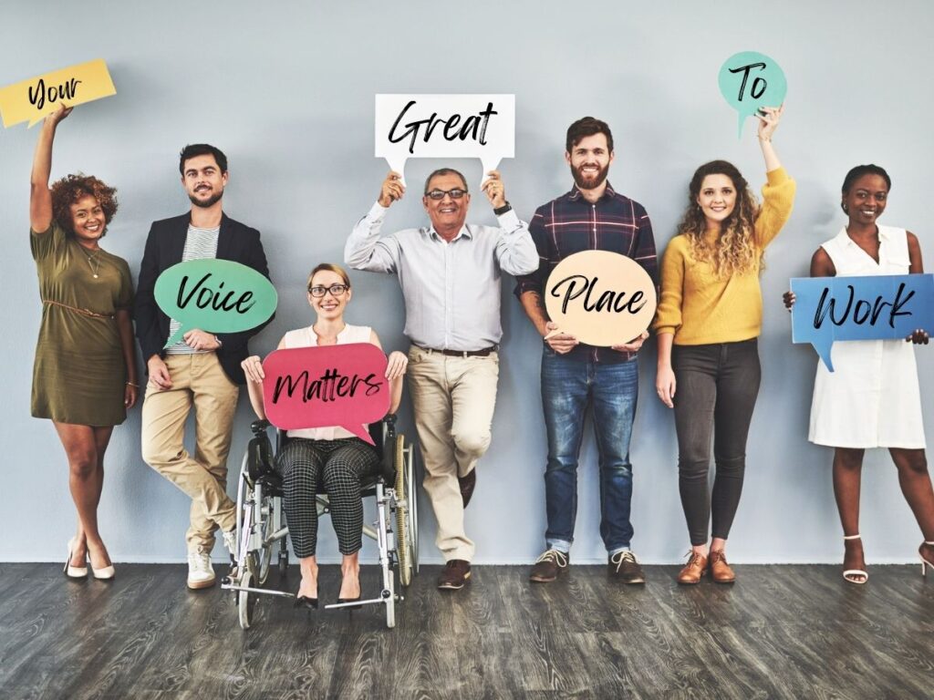 A diverse group of seven people, including a woman in a wheelchair, stand together holding colorful speech bubbles that spell out "Your Voice Matters, Great Place to Work."