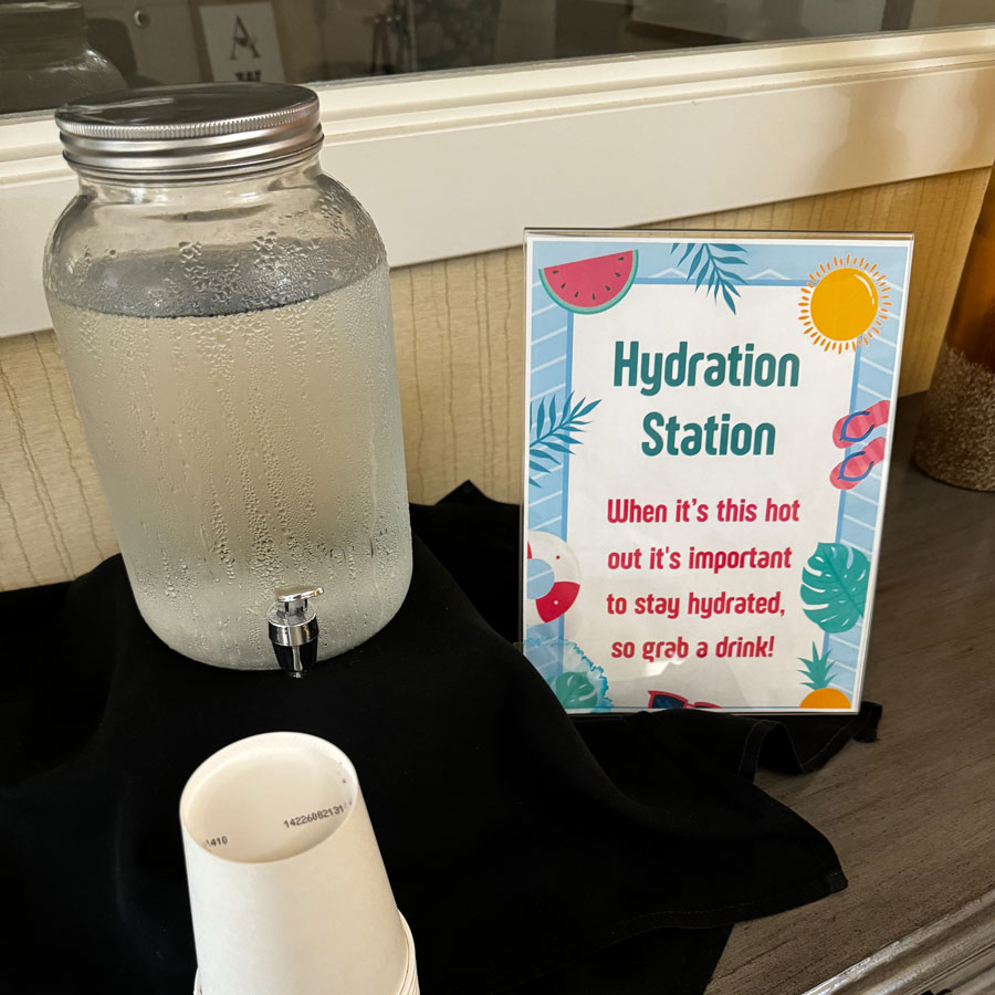 Hydration station with a large water dispenser and a sign reminding to stay hydrated in hot weather. Grab a drink!