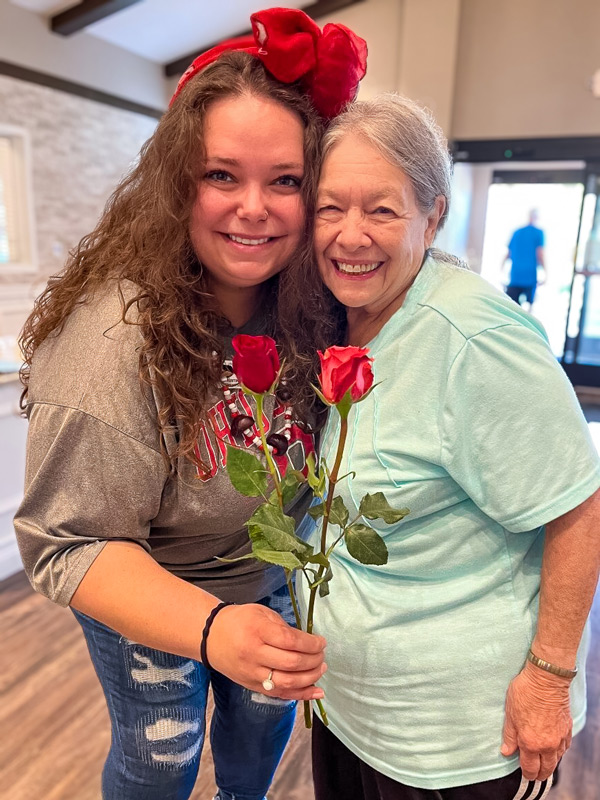 Smiling senior resident and younger woman pose together, holding two red roses. The younger woman wears a red bow and casual shirt, sharing a moment of joy in a warmly lit room.