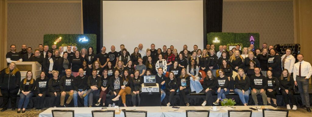 A large group photo of Arrow Senior Living team members gathered at a summit event, smiling and posing in front of a decorated stage with a positive atmosphere.